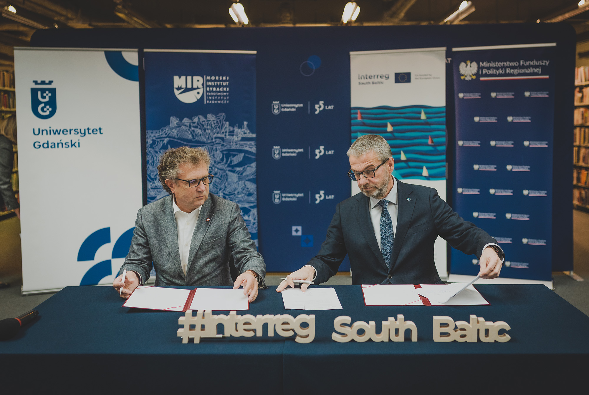 Na zdjęciu dwóch mężczyzn siedzi przy stole podczas uroczystego podpisania umowy o dofinansowanie projektu AIMused w ramach programu Interreg South Baltic. Na stole widoczny jest napis „#Interreg South Baltic”, a w tle stoją roll-upy z logotypami Morskiego Instytutu Rybackiego – PIB, Uniwersytetu Gdańskiego, Ministerstwa Funduszy i Polityki Regionalnej oraz programu Interreg South Baltic.