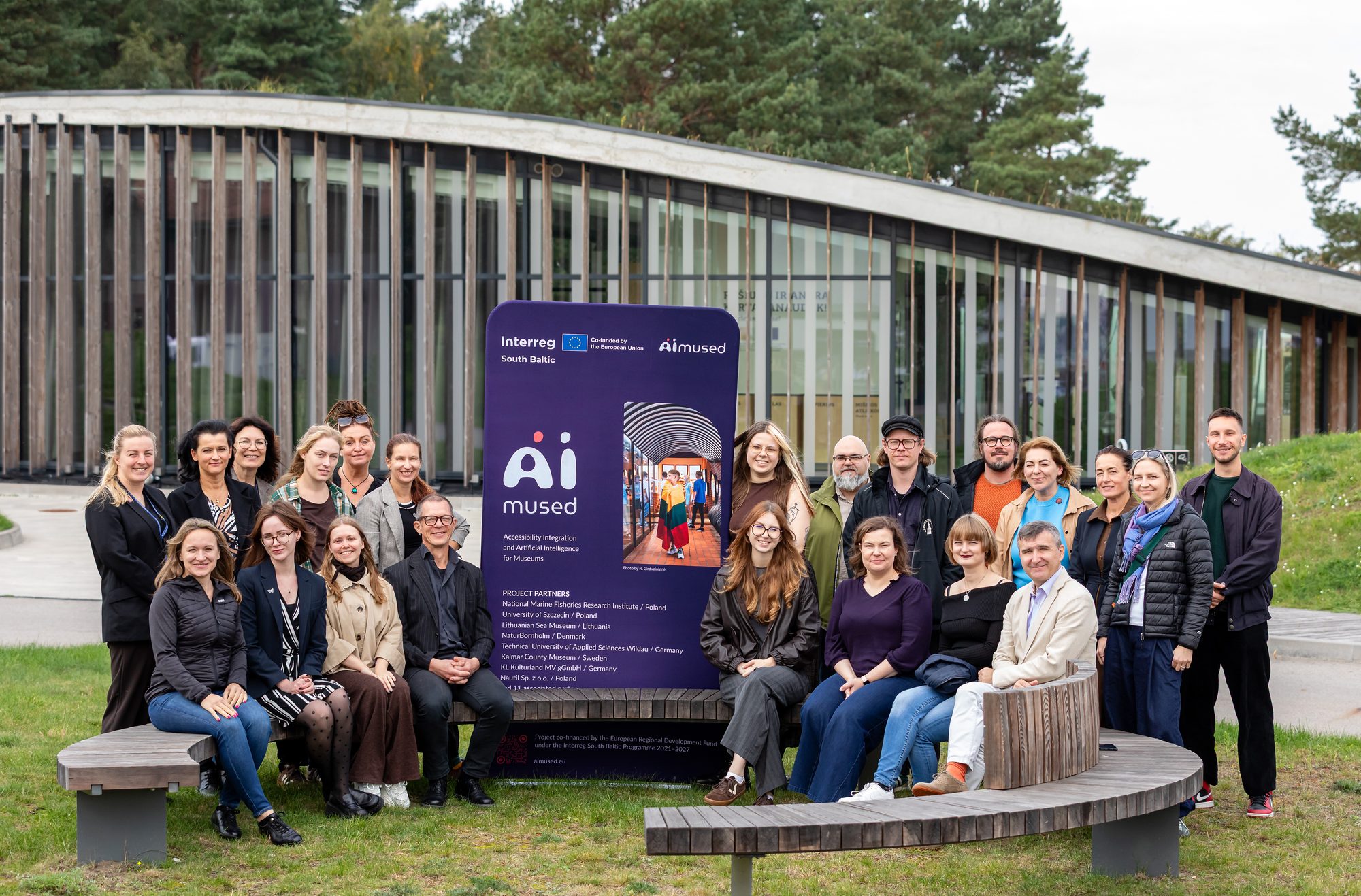 Zdjęcie grupowe partnerów projektu zgromadzonych wokół ścianki tekstylnej z logo projektu i logo programu Interreg South Baltic.